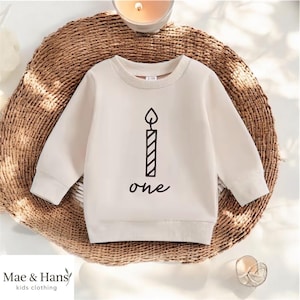 Könnte beinhalten: Cremefarbenes Langarm-Sweatshirt mit einer schwarzen Kerzengrafik und dem Wort "one" darunter. Das Sweatshirt ist für Kinder konzipiert und aus einem weichen Material gefertigt. Der Markenname "Mae & Hans kids clothing" ist unten aufgedruckt.