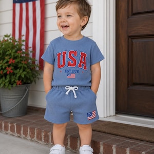 Puede incluir: Un niño con un conjunto azul de punto de gofre con la palabra "USA" en rojo en la parte superior, junto con "EST. 1776" y una pequeña bandera americana. Los pantalones cortos tienen una bandera a juego. El niño lleva zapatillas blancas.