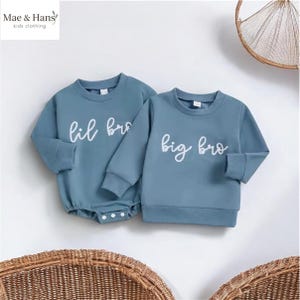 May include: Two matching blue-green baby outfits. One is a long-sleeved bodysuit with the words "lil bro" in white script. The other is a long-sleeved sweatshirt with "big bro" in white script. The brand name "Mae & Hans kids clothing" is in the top left corner.