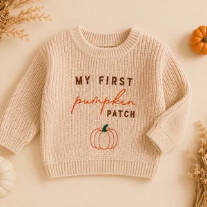 Puede incluir: Un suéter de punto beige de manga larga con el texto MY FIRST pumpkin PATCH en letras marrones y naranjas, con un gráfico de calabaza naranja bordado centrado debajo del texto en la parte delantera de la prenda.