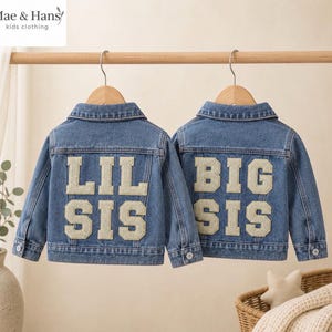以下が含まれることがあります： 木製ハンガーに掛けられた2着の青いデニムジャケット。左のジャケットの背面には白い文字でLIL SIS、右のジャケットの背面には白い文字でBIG SISと書かれています。どちらのジャケットにも襟、長袖、ボタンが付いています。