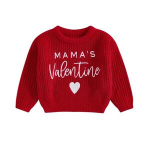 May include: A red knit sweater with long sleeves. The sweater features the text "MAMA'S Valentine" in white script, with a white heart below the text. The sweater is designed for children.