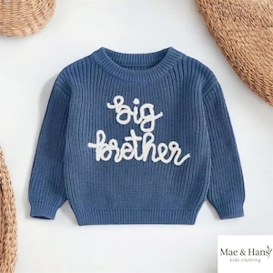 Puede incluir: Suéter de punto azul con las palabras "big brother" en escritura blanca. El suéter tiene cuello redondo y puños acanalados. El logotipo de Mae & Hans kids clothing está en la esquina inferior derecha.