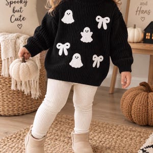 Puede incluir: Un suéter de punto negro de manga larga con motivos bordados en blanco de fantasmas y lazos, combinado con leggings acanalados de color crema y botines de ante color canela. El suéter tiene cuello redondo y una textura de punto grueso.