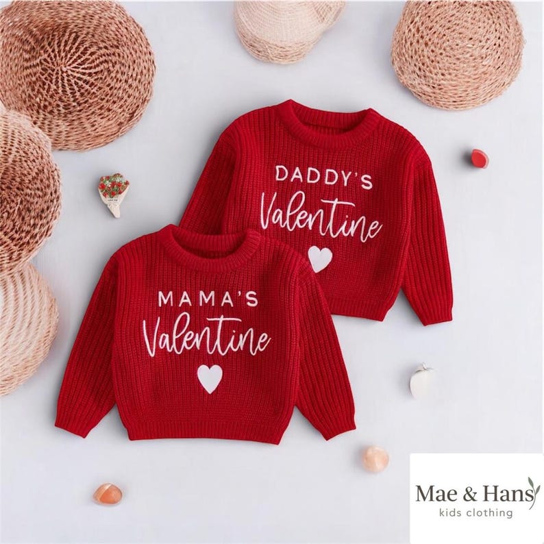 May include: Two red sweaters with white text and heart graphics. One says "MAMA'S Valentine" and the other says "DADDY'S Valentine". The sweaters are part of the "Mae & Hans kids clothing" collection.