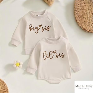 May include: Two matching cream-colored baby outfits. One is a long-sleeved sweatshirt with "big sis" in brown script. The other is a long-sleeved bodysuit with "lil sis" in brown script. The outfits are from Mae & Hans kids clothing.