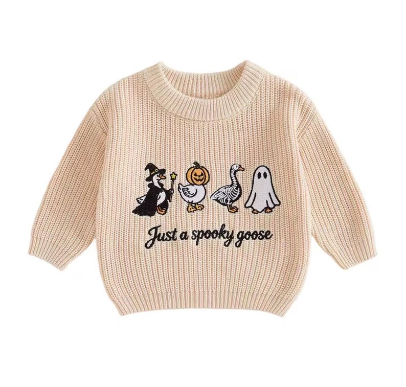 May include: A beige long-sleeve crewneck sweater featuring a ribbed knit texture. The front displays an embroidered graphic of four geese in costumes, including a witch, a pumpkin, a skeleton, and a ghost, with the text Just a spooky goose.