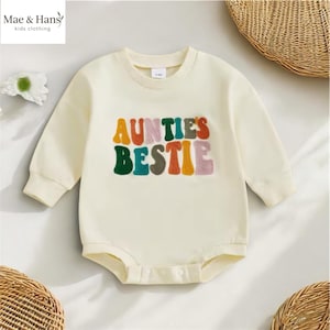May include: Cream-colored baby bodysuit with long sleeves. The front features the text "AUNTIES BESTIE" in colorful, retro-style lettering. The bodysuit is made of soft material and has a round neckline. The brand name "Mae & Hans kids clothing" is in the top left corner.