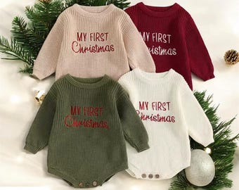 May include: Four baby Christmas sweaters in beige, burgundy, olive green, and white. Each sweater has long sleeves and the embroidered text "MY FIRST Christmas" in red. The sweaters are knit and have button closures at the bottom.