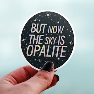 May include: A round sticker with the text "BUT NOW THE SKY IS OPALITE" in a holographic font. The sticker has a black background with white stars and a white border. The sticker is held in a hand.