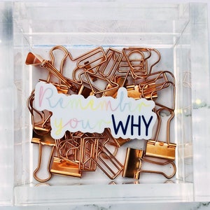 May include: A clear acrylic box filled with rose gold binder clips and a sticker that reads "Remember your WHY" in rainbow colors.