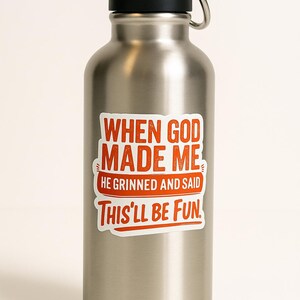 May include: A stainless steel water bottle with a black handle and lid. The bottle features a sticker with the text "WHEN GOD MADE ME HE GRINNED AND SAID THIS'LL BE FUN" in red and white.