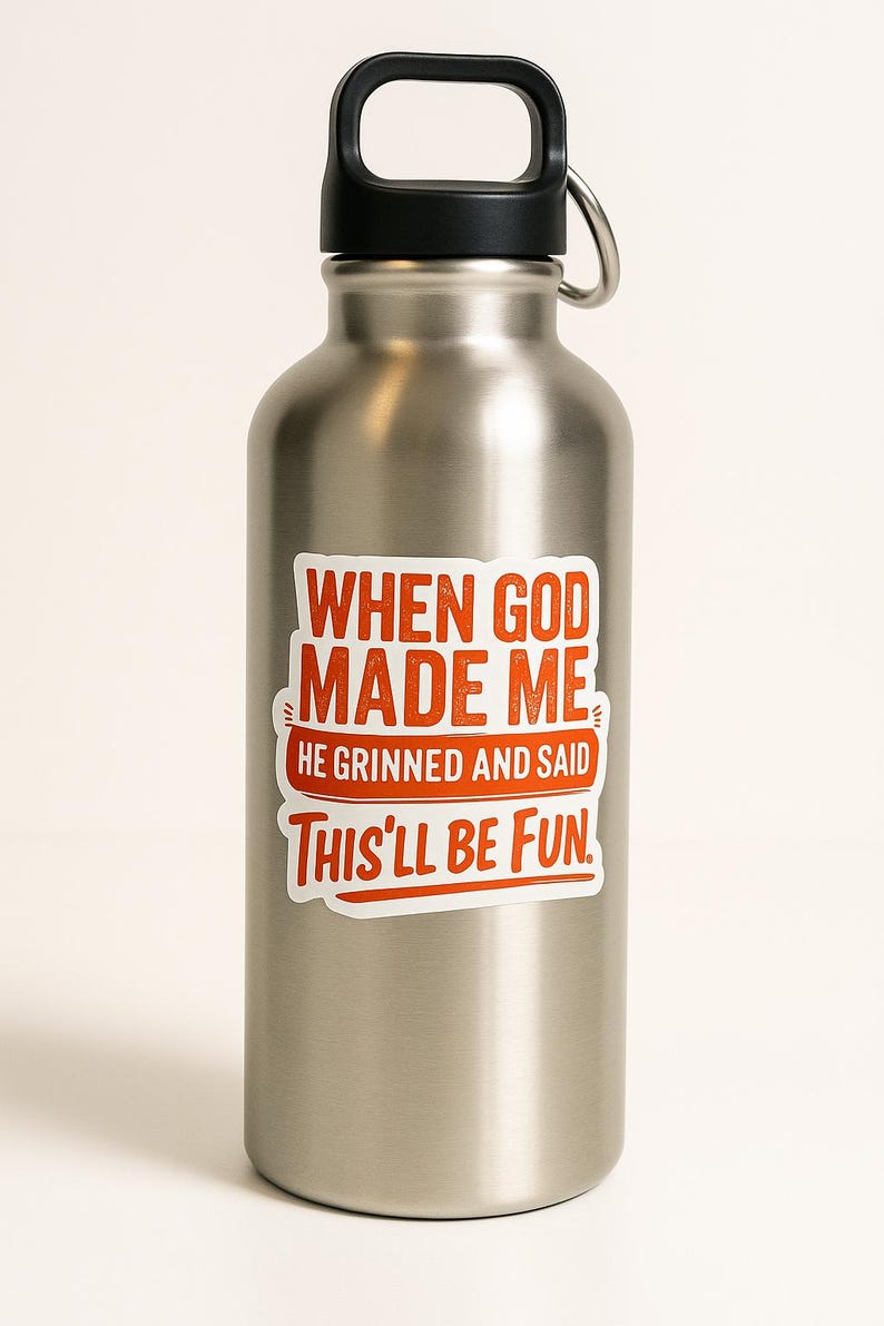 May include: A stainless steel water bottle with a black handle and lid. The bottle features a sticker with the text "WHEN GOD MADE ME HE GRINNED AND SAID THIS'LL BE FUN" in red and white.