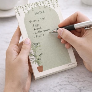 May include: A notepad with a green and beige design, featuring a grocery list with items like eggs and bread. The notepad has a botanical illustration and the word "NOTES" at the top. A hand is writing on the notepad with a pen.