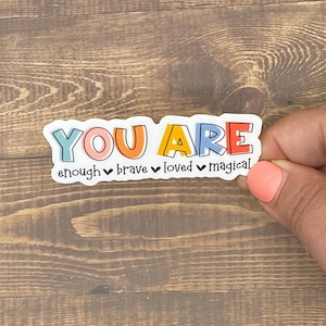 May include: A colorful sticker with the words "YOU ARE" in large letters. Below the words are the words "enough, brave, loved, magical" with a heart symbol between each word.