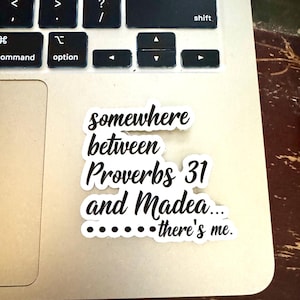 Puede incluir: Una pegatina blanca con texto negro que dice "en algún lugar entre Proverbios 31 y Madea... ahí estoy yo". La pegatina está sobre el teclado de un portátil.
