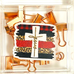 May include: A clear acrylic box filled with gold binder clips and a sticker with a cross and the word "Jesus" on it. The sticker has a red, black, and white plaid pattern, a leopard print, and red glitter.
