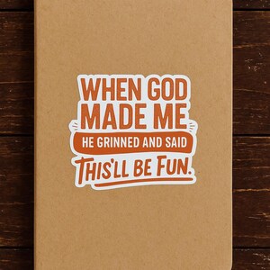 May include: A tan notebook with rounded corners. A sticker with the text "WHEN GOD MADE ME, HE GRINNED AND SAID THIS'LL BE FUN" is affixed to the front. The text is in orange and white.