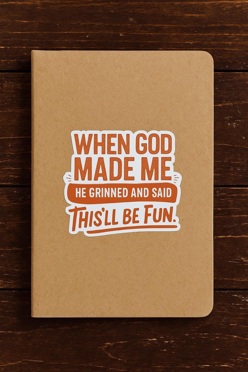 May include: A tan notebook with rounded corners. A sticker with the text "WHEN GOD MADE ME, HE GRINNED AND SAID THIS'LL BE FUN" is affixed to the front. The text is in orange and white.