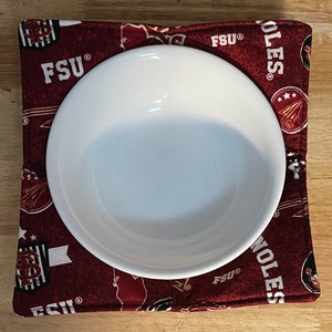 May include: A red and white fabric hot pad with the text "FSU" and "NOLES" printed on it. A white bowl is sitting on top of the hot pad.