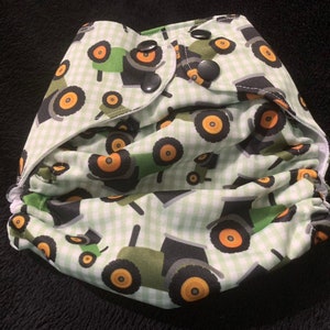 May include: A green and white cloth diaper with a tractor print. The diaper has a white gingham pattern and features multiple green tractors with orange wheels.