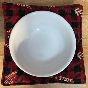 May include: A red and black checked fabric hot pad with the words "Arizona State" in gold lettering. A white bowl is sitting on top of the hot pad.