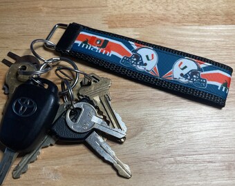 May include: A black key chain with a Miami Dolphins themed fabric strap. The strap has a white, orange, and blue design with the team logo and the word "Miami". The keychain is attached to a set of keys, including a car key with a black fob.