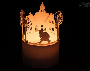 Może przedstawiać: Papierowy diorama przedstawiający małe miasteczko z kościołem, drzewami i sylwetką osoby idącej. Diorama jest podświetlone od wewnątrz, tworząc ciepłe światło.