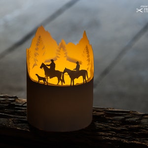 Könnte beinhalten: Eine weiße Papierlaterne mit einer Silhouette von zwei Personen, die auf Pferden reiten, und einem Hund in einer Waldlandschaft. Die Laterne wird von innen beleuchtet und strahlt ein warmes Licht aus.