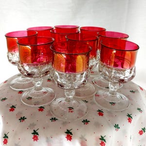 Peut inclure: Ensemble de dix verres à vin à pied en verre transparent avec une finition rouge irisée. Les verres sont disposés en cercle sur une nappe blanche à motif floral rouge et vert.