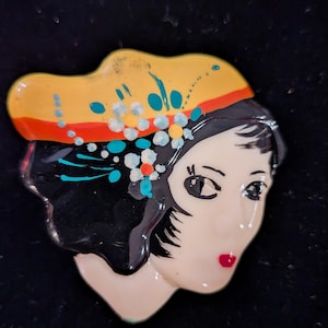 May include: A glossy brooch featuring the stylized face of a person with black hair, red lips, and dark eyes. The figure wears a yellow and orange headpiece adorned with small white and teal floral accents, all rendered in a smooth, reflective finish.