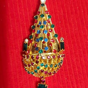 Peut inclure: Une broche dorée ornée en forme de lustre, incrustée de pierres précieuses rouges, vertes et bleues. La broche a une forme de goutte d'eau avec une pierre verte rectangulaire en bas et une gemme transparente en haut. Le fond est rouge uni.