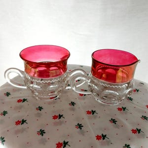 Puede incluir: Un juego de dos jarras de crema y azúcar de vidrio transparente con bordes de vidrio rojo. Las jarras tienen un diseño de vidrio cortado y están sobre un mantel blanco con un patrón floral rojo y verde.