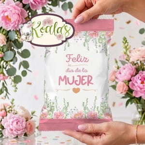 Puede incluir: Una bolsa blanca con un diseño floral rosa y las palabras "Feliz día de la MUJER" en rosa. La bolsa tiene un logotipo de Koala's y está sostenida por una persona. El fondo presenta flores rosas y vegetación.