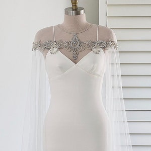 May include: A white wedding dress with a tulle cape featuring a beaded neckline. The dress has a fitted bodice and a flowing skirt.