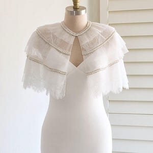 May include: A white wedding dress with a long, flowing skirt and a fitted bodice. The dress features a delicate lace overlay and a pearl-embellished cape.