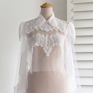 May include: A white lace bridal bolero with a high collar and long sleeves. The bolero is displayed on a wooden mannequin stand against a white background.