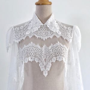 May include: White lace bridal bolero with a high collar and long sleeves. The bolero is made of delicate lace and has a sheer, flowing design.