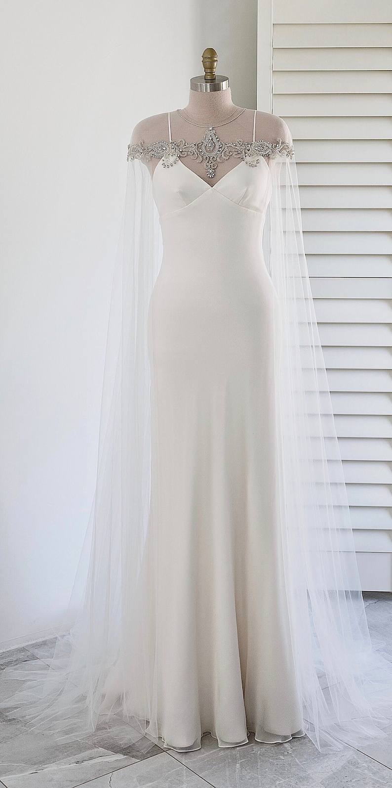 May include: A white wedding dress with a tulle cape featuring a beaded neckline. The dress has a fitted bodice and a flowing skirt.