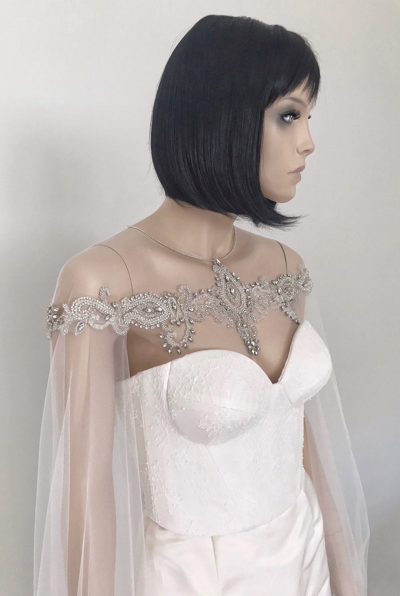 May include: A white bridal cape with a beaded and sequined lace design. The cape is sheer and has a delicate, feminine look.