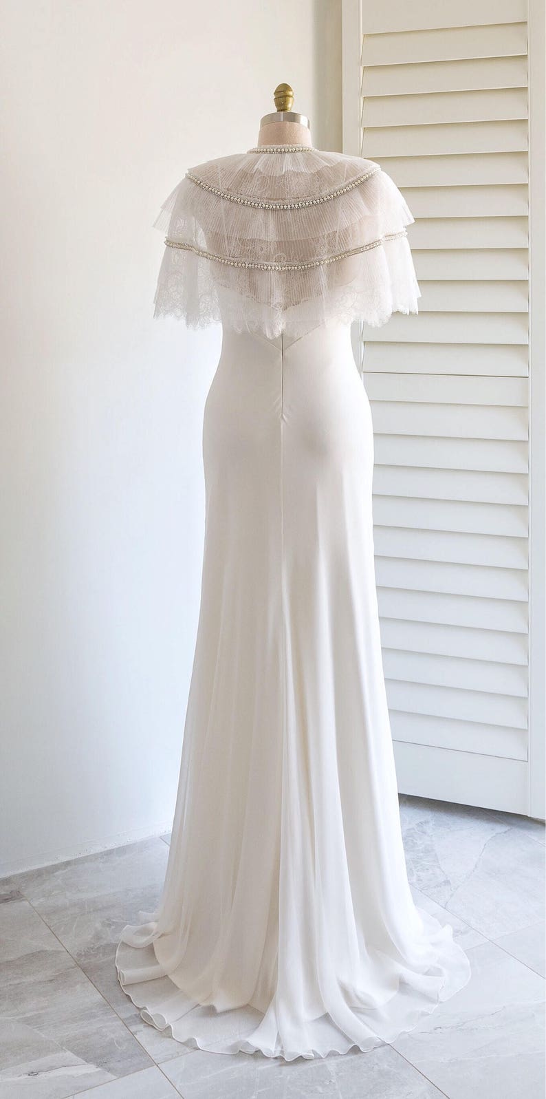 May include: A white wedding dress with a long, flowing skirt and a lace bodice. The dress has a sheer, layered cape with pearl embellishments.