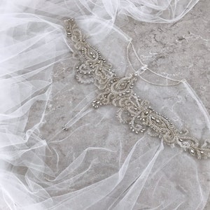 May include: A delicate white tulle veil with a beaded embellishment featuring a floral design. The embellishment is made of silver beads and rhinestones.