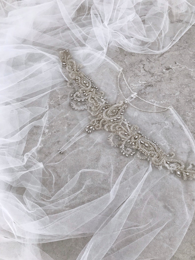 May include: A delicate white tulle veil with a beaded embellishment featuring a floral design. The embellishment is made of silver beads and rhinestones.