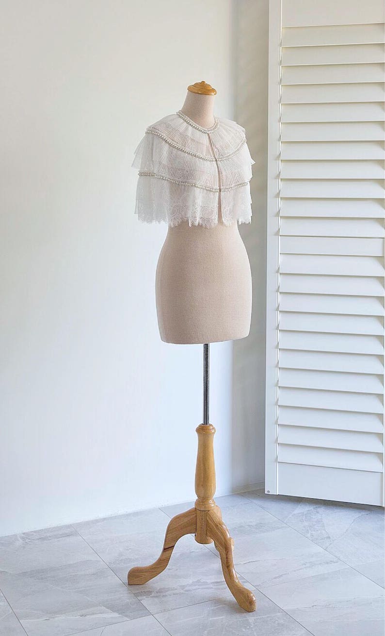 May include: A white lace bridal cape with pearl embellishments, displayed on a wooden mannequin stand.