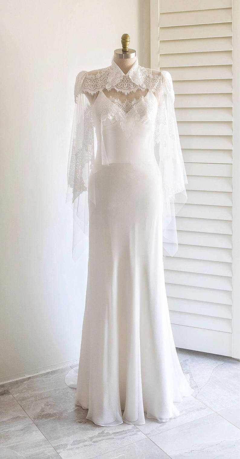 May include: A white wedding dress with a lace overlay and a high neckline. The dress has a fitted bodice and a flowing skirt.