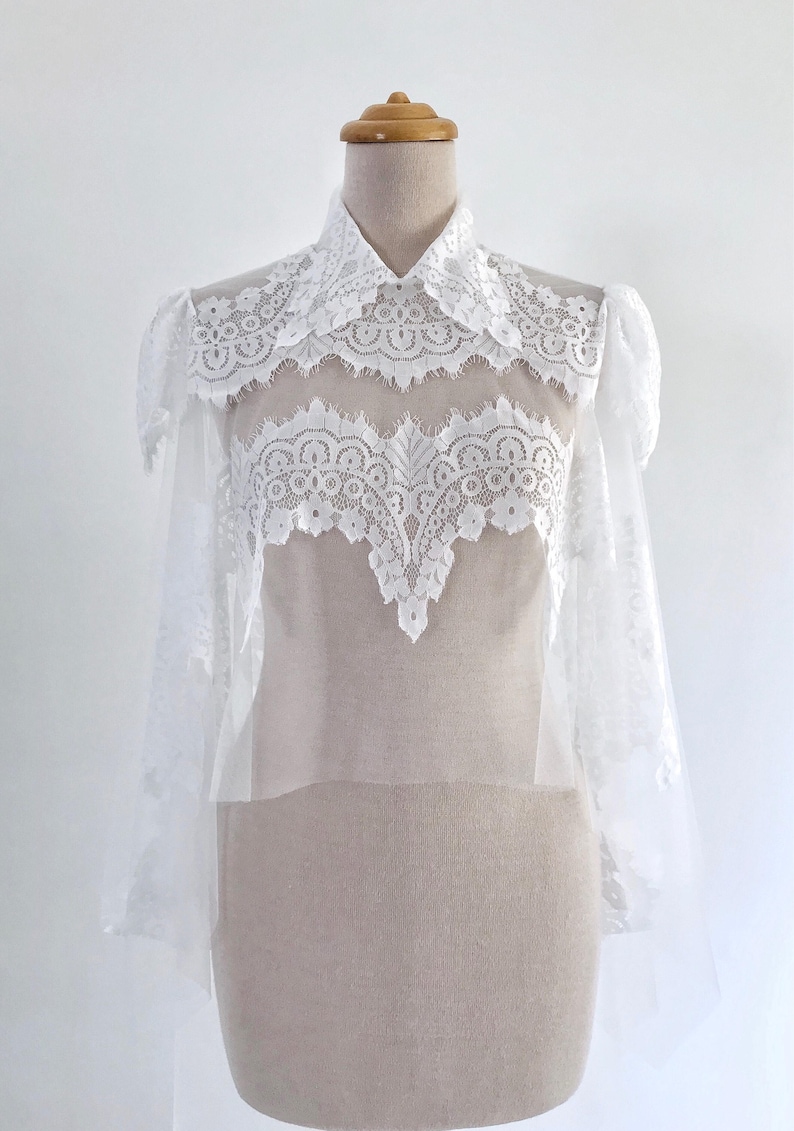 May include: White lace bridal bolero with a high collar and long sleeves. The bolero is made of delicate lace and has a sheer, flowing design.