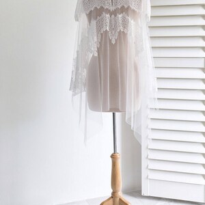 May include: White lace bridal cape with a high neck and long sleeves. The cape is made of delicate tulle and features intricate lace detailing. The cape is displayed on a wooden mannequin.