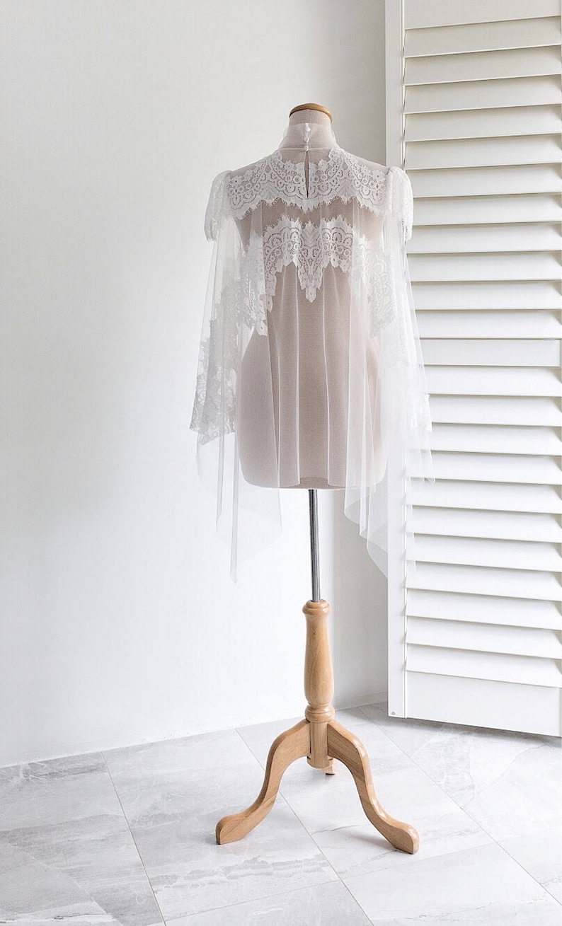 May include: White lace bridal cape with a high neck and long sleeves. The cape is made of delicate tulle and features intricate lace detailing. The cape is displayed on a wooden mannequin.