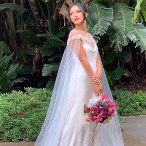 May include: A white wedding dress with a tulle cape and a floral bouquet. The dress has a fitted bodice and a flowing skirt. The bride is standing in front of a lush green background.