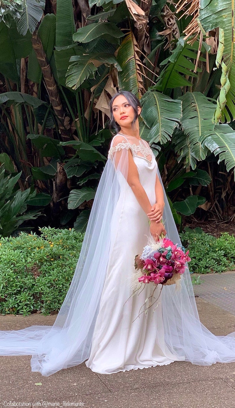 May include: A white wedding dress with a tulle cape and a floral bouquet. The dress has a fitted bodice and a flowing skirt. The bride is standing in front of a lush green background.